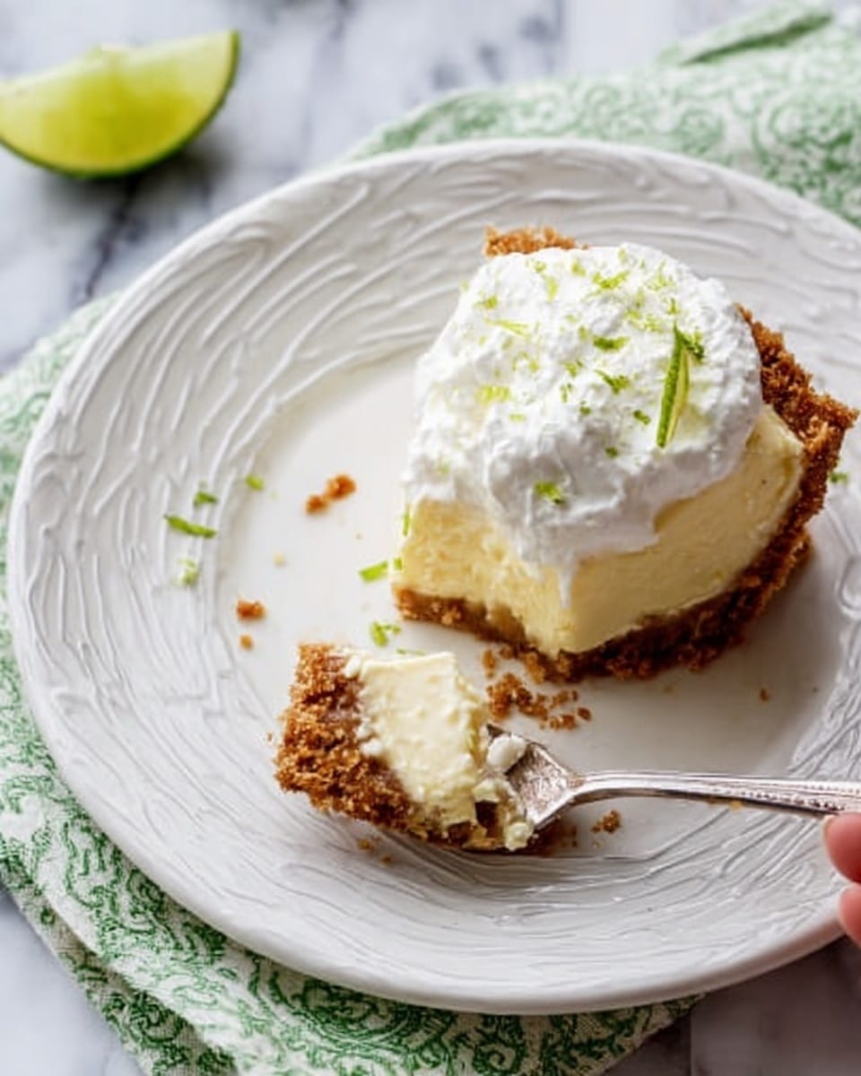 A glass pie dish contains a three-layer dessert with a crumbly golden brown crust forming the base and sides. The middle layer is smooth, pale yellow with small green lime zest pieces scattered on top. Around the edge of the dessert, there are evenly spaced dollops of white whipped cream, each with a soft, swirled texture. This is set on a white marbled surface, and a silver utensil with a worn handle lies nearby on the right. photo taken with an iphone --ar 4:5 --v 7