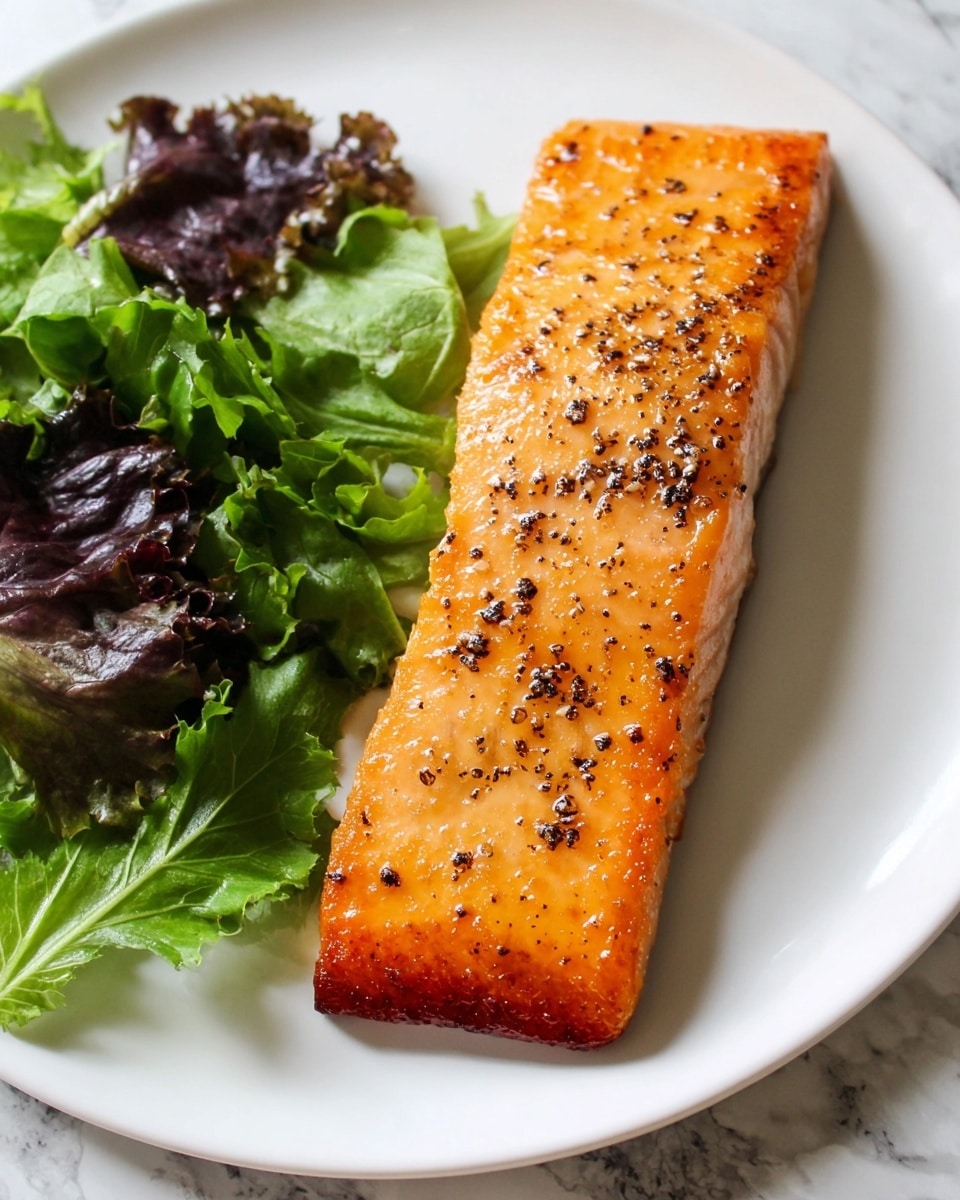 A piece of cooked salmon with a shiny orange top layer covered in black pepper sits in the center of a white plate, showing slightly crispy brown edges. To the right side of the salmon, there is a small pile of fresh mixed green and purple lettuce leaves with a leafy texture. The plate rests on a white marbled surface. photo taken with an iphone --ar 4:5 --v 7