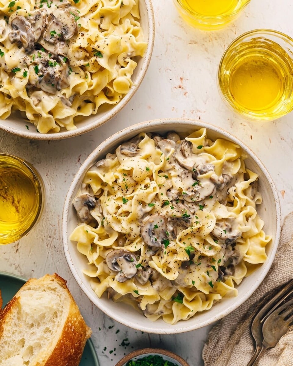 Two white bowls filled with creamy pasta layered with small curls of wide egg noodles mixed evenly with chopped mushrooms coated in a white, smooth sauce, garnished lightly with chopped green herbs and a sprinkle of black pepper on top. Beside the bowls are two clear glasses filled with a yellow liquid, placed on a white marbled surface. There are pieces of bread with a soft texture near the bowls, and a small plate with green herbs is partially visible in the corner. The overall image is bright and natural with focus on the creamy, textured pasta. photo taken with an iphone --ar 4:5 --v 7