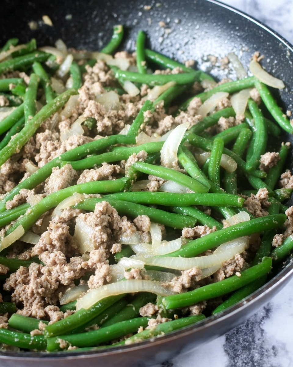 The image shows a pan with cooked green beans mixed with ground light brown meat and white onion slices. The green beans are long and bright green, lying on top of the crumbled meat with soft, slightly translucent onion pieces spread evenly throughout. The mixture looks moist and well-cooked, resting on a dark pan surface with some light reflections. The background has a white marbled texture. Photo taken with an iphone --ar 4:5 --v 7