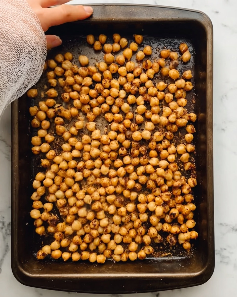The image shows a single layer of golden-brown roasted chickpeas spread evenly in a dark baking tray. The chickpeas have a slightly rough texture with small charred spots and a light coating of oil, showing a mix of warm tan and amber colors. A white mesh cloth is partially visible in the top left corner, with a woman’s hand gently touching the chickpeas. The tray is sitting on a white marbled surface. photo taken with an iphone --ar 4:5 --v 7