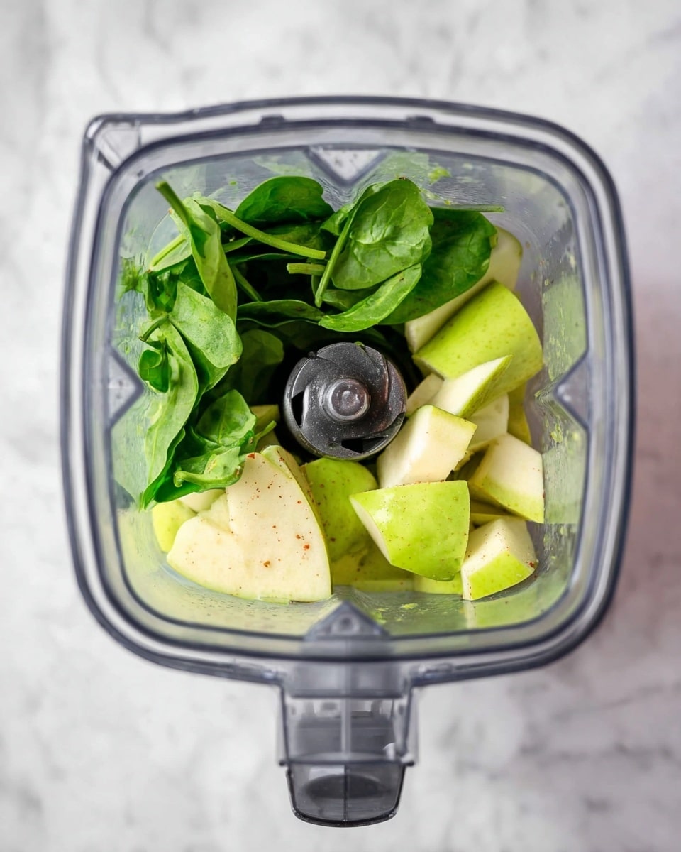 The image shows a top view of a blender container filled with ingredients for a smoothie. Inside, there are fresh green spinach leaves at the bottom, layered with chunks of light green apple pieces on top. The blender blades are visible in the center with the clear container edges surrounding the contents. The background surface has a white marbled texture photo taken with an iphone --ar 4:5 --v 7