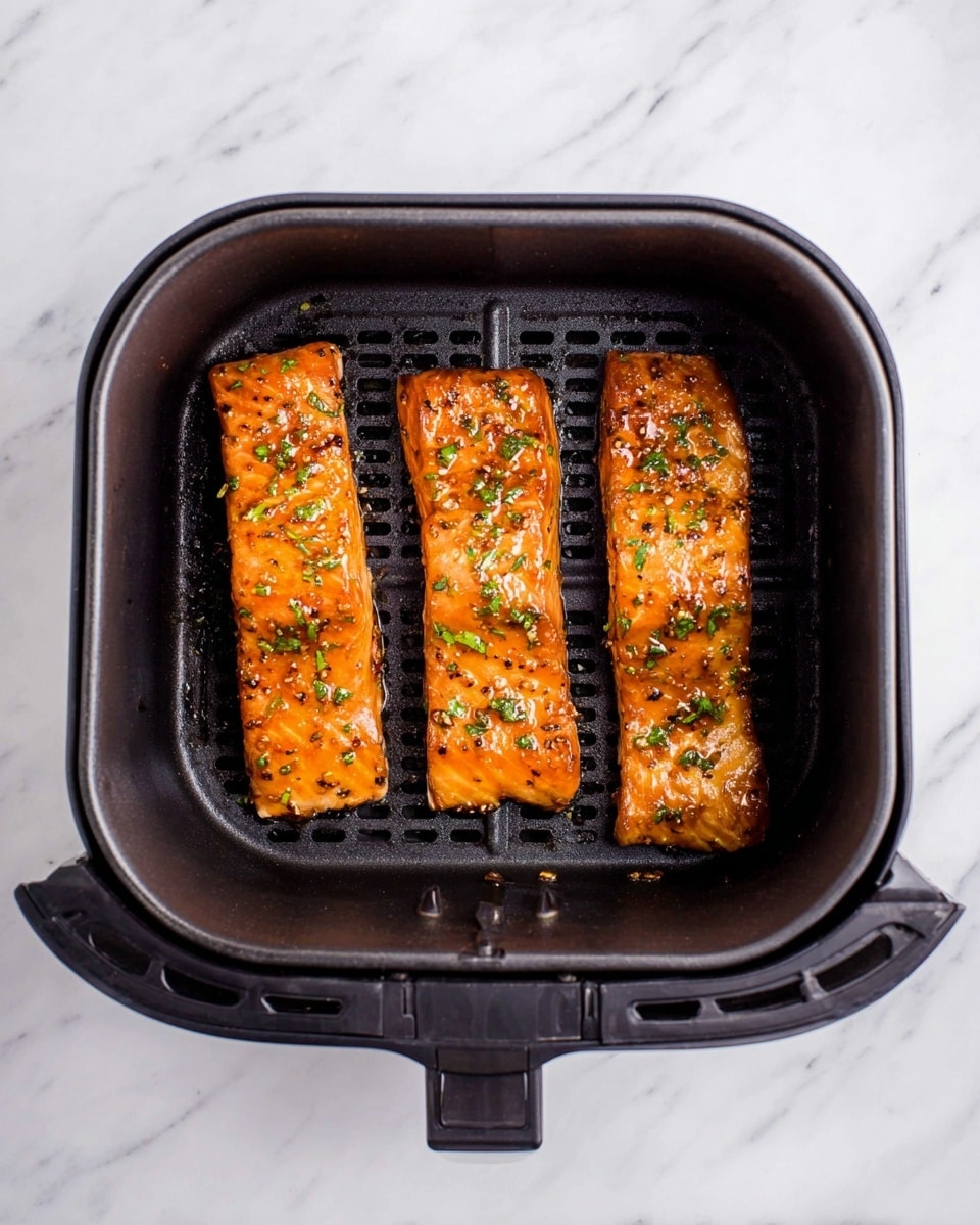 Three pieces of cooked salmon with a shiny orange glaze lay side by side inside a black air fryer basket. Each salmon piece has bits of green herbs scattered on top, adding a touch of color to the smooth, slightly crispy surface. The basket has a grid pattern beneath the fish, and the whole setup sits on a white marbled surface. photo taken with an iphone --ar 4:5 --v 7
