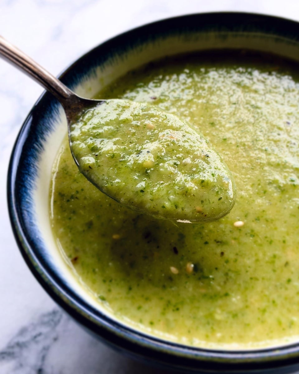 A close-up view of a green sauce or soup with a thick, slightly chunky texture containing visible small seeds. The sauce fills a white bowl with a dark blue interior rim. A metal spoon is partially dipped into the sauce, lifting a scoop that shows the texture clearly. The background is a white marbled surface. photo taken with an iphone --ar 4:5 --v 7