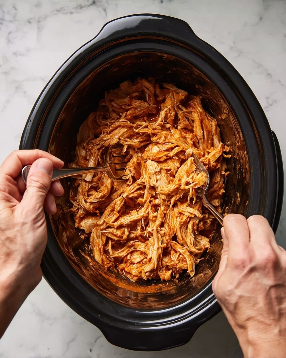 A close-up top view shows shredded orange-colored chicken inside a black slow cooker. Two adult hands, one on each side, hold forks and pull the chicken apart, creating a mix of stringy and chunky textures throughout the meat. The slow cooker sits on a surface with a white marbled texture. The contrast between the cooked chicken and the dark black pot is clear, highlighting the rich, warm colors of the meat. photo taken with an iphone --ar 4:5 --v 7