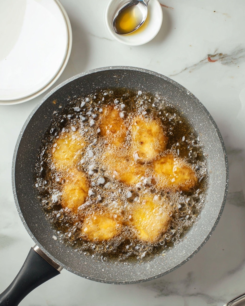 A round gray pan with a speckled pattern is shown from above on a white marbled surface. Inside the pan, several golden-yellow pieces of food are frying in bubbling hot oil, creating many small, shiny bubbles that cover the surface of the oil. The pan handle is black and angled to the side. Nearby, there are white dishes, one of which holds a spoon with some light sauce on it. Photo taken with an iphone --ar 4:5 --v 7