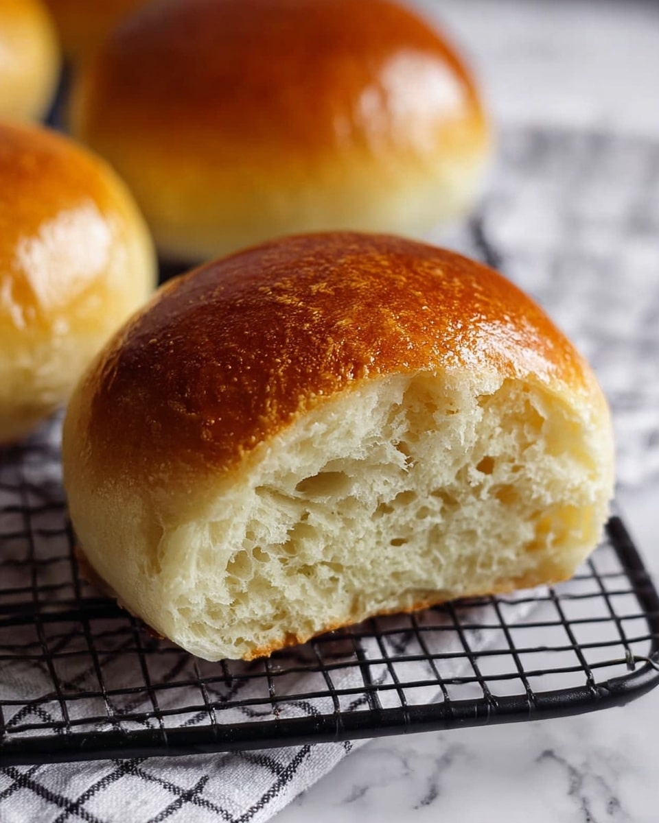 A close-up view of a soft round bread bun split in half, showing a golden brown top layer with a shiny and smooth texture, and a light creamy inside with small air holes and a fluffy, soft look. The bun rests on a black wire cooling rack, which sits on a white marbled surface with a black and white grid-patterned cloth underneath. In the background, there are slightly blurred similar buns, giving a warm and fresh baked feeling. photo taken with an iphone --ar 4:5 --v 7