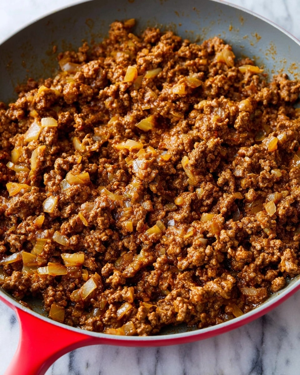 A close-up view of a red skillet filled with cooked ground beef mixed with small pieces of cooked onions, all showing a rich brown and golden color from cooking. The texture looks soft and slightly crumbly with bits of onion evenly spread throughout the meat. The skillet is placed on a white marbled surface. photo taken with an iphone --ar 4:5 --v 7