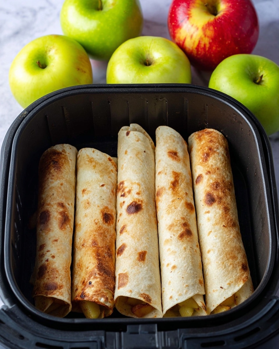 Five rolled light brown baked tortillas with some browned spots lay side by side inside a black air fryer basket, with some golden cooked apple pieces slightly visible at the ends of a few tortillas. In the background, five whole apples with shiny skins are placed on a white marbled surface—three are bright green and two are red with yellow streaks. The scene is lit softly, highlighting the textures on the tortillas and apples. Photo taken with an iphone --ar 4:5 --v 7