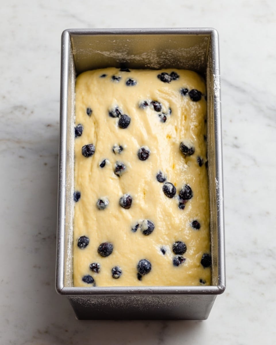 A rectangular metal baking pan filled with pale yellow batter that has small dark blueberries scattered throughout. The batter is spread evenly but with some soft, smooth ridges on the surface. The pan sits on a white marbled surface, and there is a light dusting of flour on the edges of the pan. photo taken with an iphone --ar 4:5 --v 7