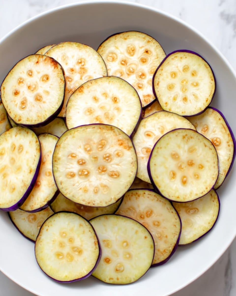 A white bowl filled with multiple round slices of eggplant stacked loosely inside. Each slice has a pale yellow center with small clusters of light brown seeds and a thin purple edge. The texture looks smooth with slight browning on some slices, and the bowl sits on a white marbled surface. photo taken with an iphone --ar 4:5 --v 7