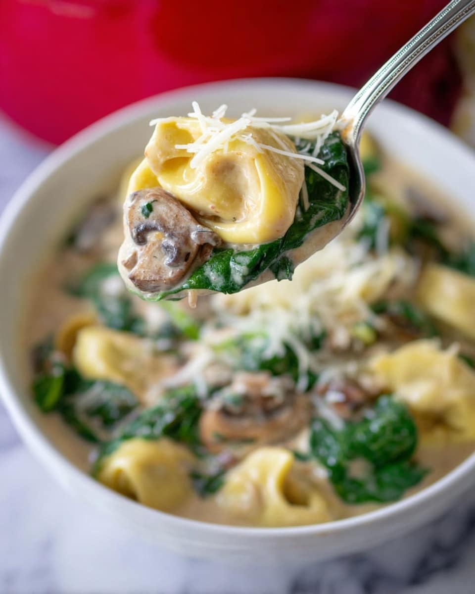 A close-up view of a silver spoon holding one piece of yellow tortellini pasta filled and folded, topped with thin strands of white shredded cheese and resting on a bed of deep green spinach leaves and small brown mushroom slices, all coated in a creamy light beige sauce. Below the spoon, a white bowl filled with more tortellini, spinach, mushrooms, and sauce is partially visible against a white marbled surface. In the background, a bright red object is slightly blurred. Photo taken with an iphone --ar 4:5 --v 7