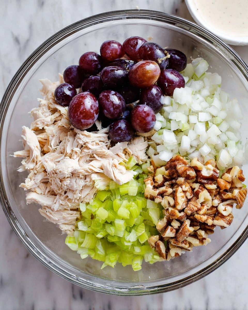 A clear glass bowl sits on a white marbled surface, filled with four main layers arranged separately. The bottom layer shows shredded pale beige chicken pieces scattered unevenly under the other layers. On the top left, half-cut dark purple grapes with shiny skins create a glossy cluster. To the top right, there are small, finely diced white onions forming a soft, textured pile. Below the onions, on the bottom right, there are light brown chopped nuts with rough and uneven shapes. On the bottom left, finely chopped celery with a fresh green color and small pieces completes the mix. A small white ceramic bowl with a creamy sauce is visible in the upper part of the image. Photo taken with an iphone --ar 4:5 --v 7