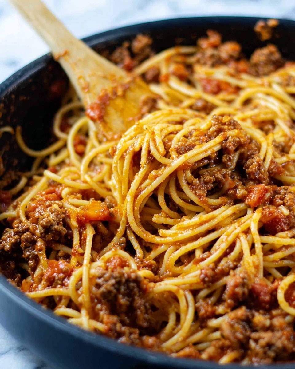 The image shows a close-up of spaghetti mixed with a thick, chunky meat sauce inside a black pan. The spaghetti is a pale yellow color, twisted around and coated in the reddish-brown meat sauce filled with small pieces of cooked ground meat and bits of tomato. A wooden spoon with some sauce on it rests in the pan behind the pasta. The background is a soft focus white marbled texture. photo taken with an iphone --ar 4:5 --v 7