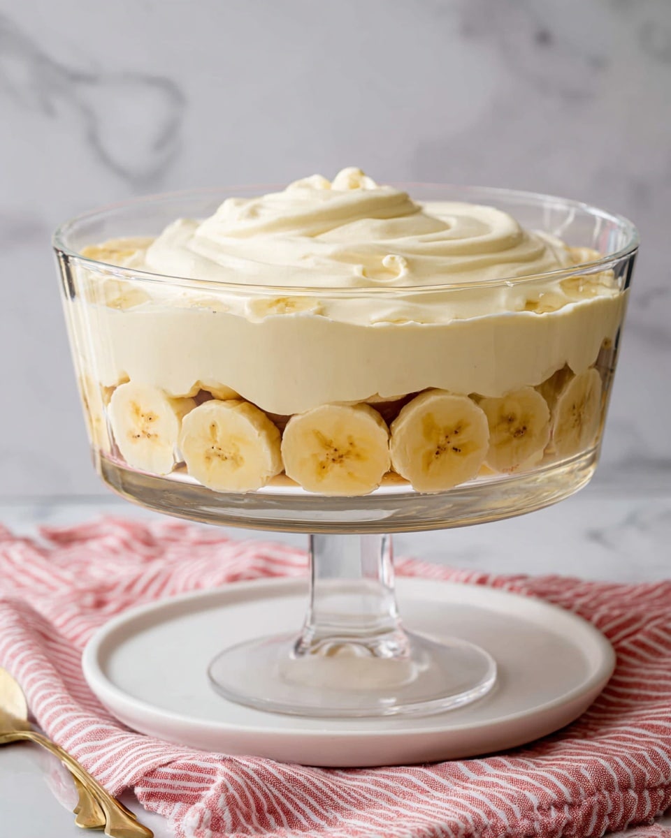 A clear glass trifle bowl on a white plate holds a dessert with two layers. The bottom layer shows round banana slices lined up around the edge, light yellow with small brown spots. Above the bananas is a thick, smooth cream layer, pale vanilla color, spread evenly and swirled on top. The bowl sits on a white marbled surface with a soft pink and white striped cloth underneath. Photo taken with an iphone --ar 4:5 --v 7