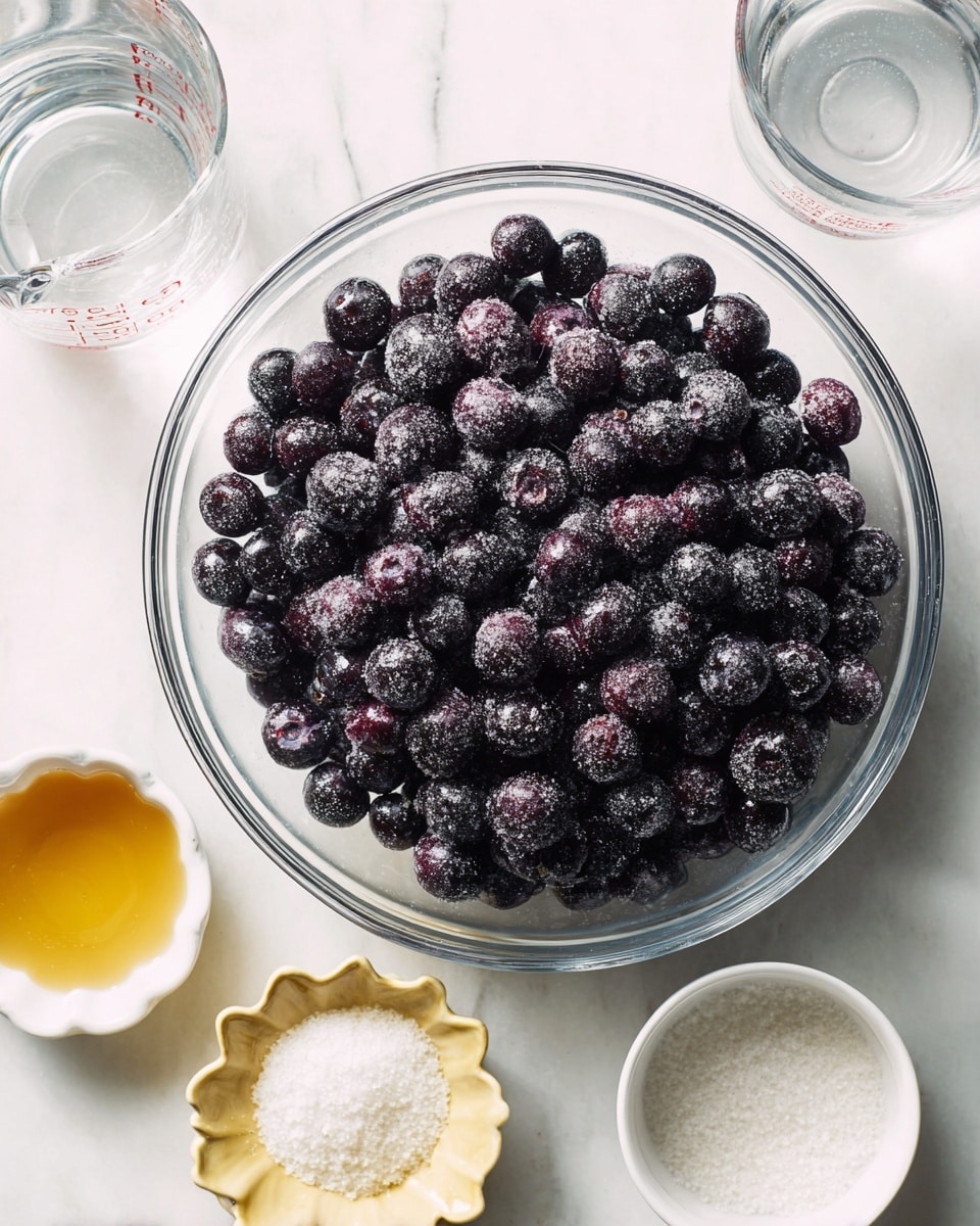 The image shows a large clear glass bowl filled with dark purple frozen blueberries, covered lightly with frost. Around the bowl, there are small white dishes with different ingredients: a scalloped dish with a golden liquid, a small bowl filled with white powder, and a round bowl with granulated white sugar. Above these, a glass measuring cup with water is partly visible. All items are set on a white marbled surface, creating a clean and fresh look. photo taken with an iphone --ar 4:5 --v 7