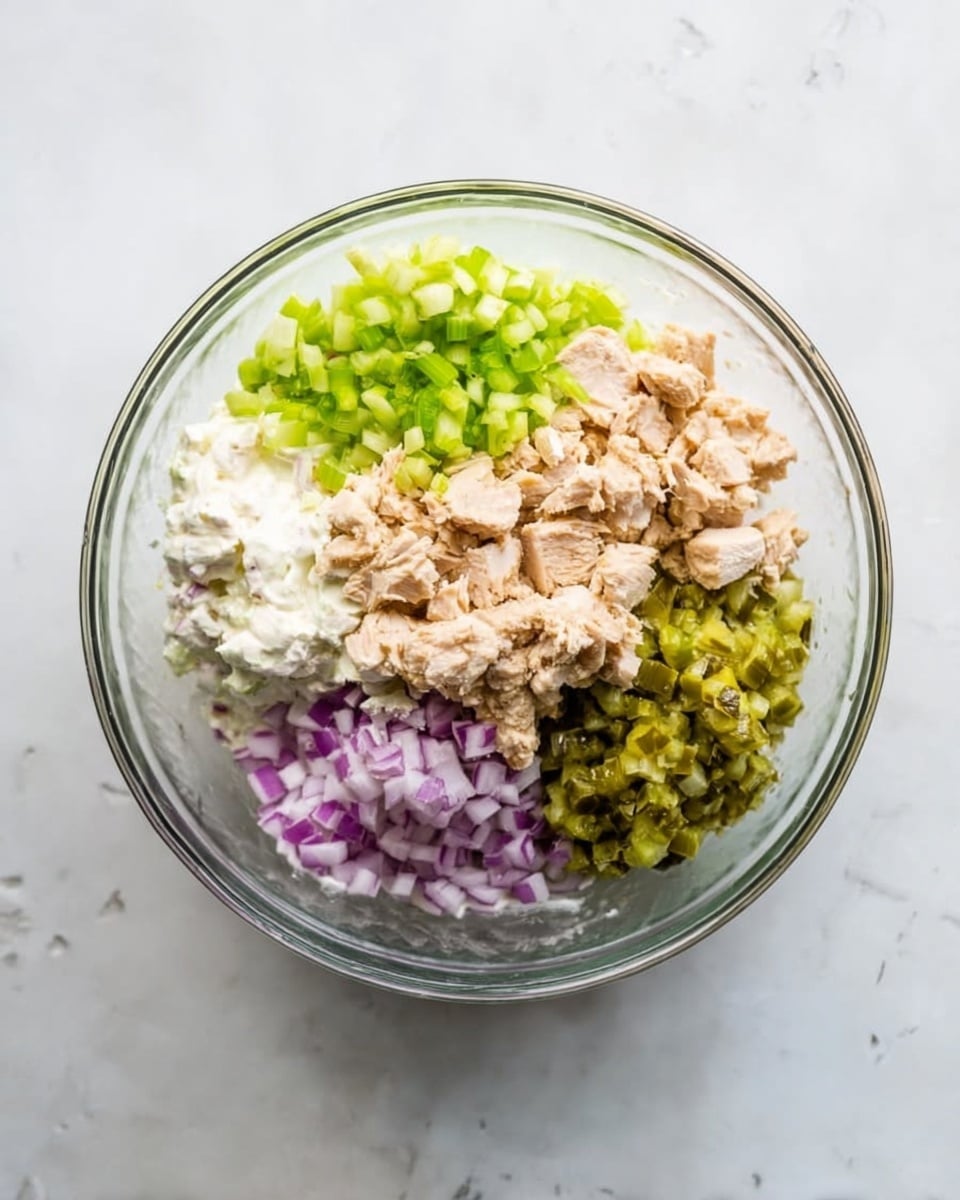 A clear glass bowl sits on a white marbled surface, with five separate layers of ingredients inside. Starting from the top left and moving clockwise, there is a bright green layer of chopped celery, a creamy white layer of mayonnaise, a chunky light brown layer of cooked chicken pieces, a vivid green layer of chopped pickles, and a finely chopped purple layer of red onions. Each layer is distinct and evenly placed, showing different colors and textures clearly. Photo taken with an iphone --ar 4:5 --v 7