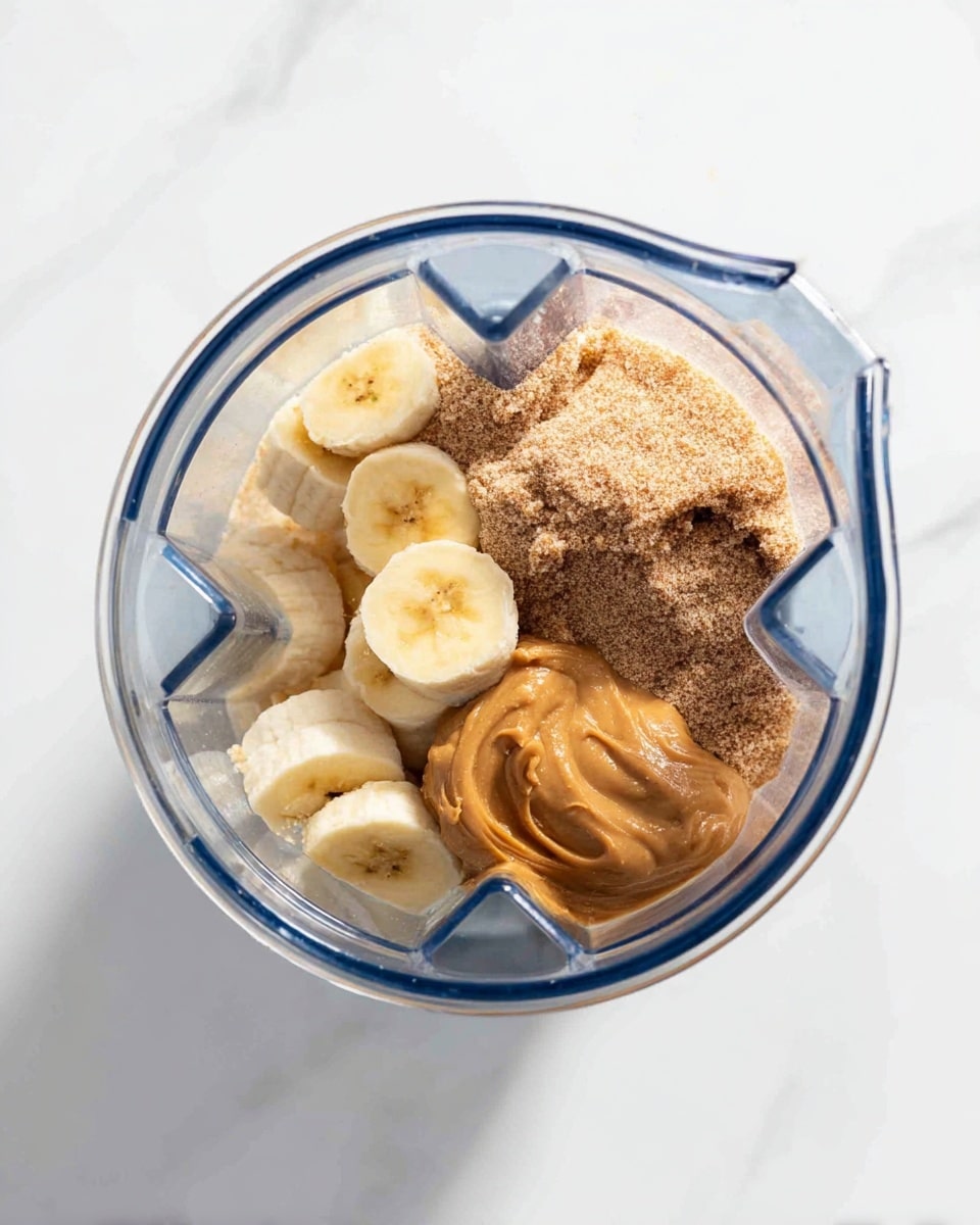 The image shows a blender container seen from above, placed on a white marbled surface. Inside the blender, there are four main layers: at the bottom, frozen banana slices, creamy light yellow with frost on their edges; next to the bananas, a light brown powder, coarse and grainy in texture; on one side, a thick dollop of peanut butter in a darker golden brown color with smooth swirls; the rest of the container is clear and transparent. The blender container has a blue rim, and the whole scene is lit brightly with clear focus. photo taken with an iphone --ar 4:5 --v 7