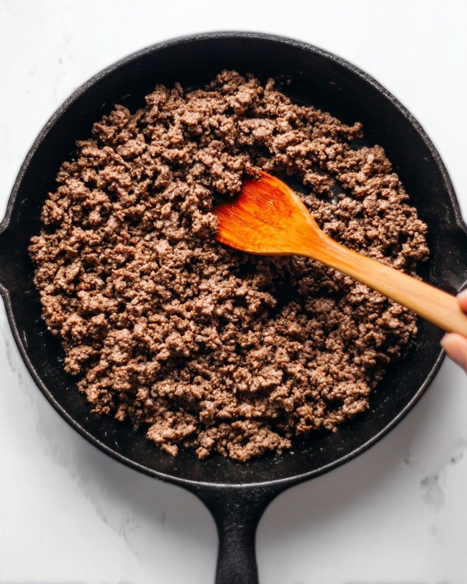 The image shows a black cast iron pan filled with cooked ground beef. The beef is brown and crumbly, spread evenly in one layer inside the pan. A woman's hand is holding a wooden spoon, stirring the beef. The background is a white marbled surface. photo taken with an iphone --ar 4:5 --v 7