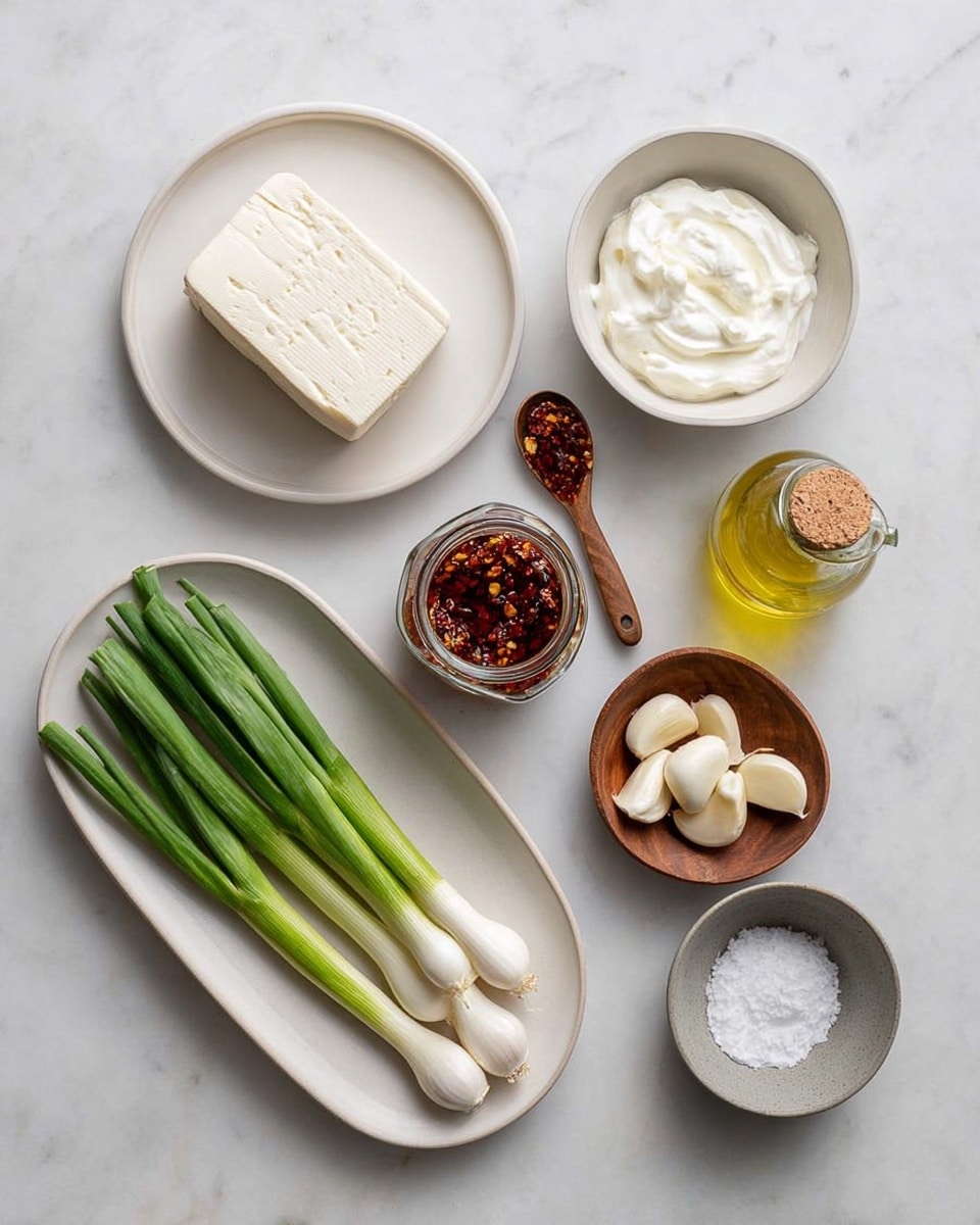 The image shows seven ingredients placed neatly on a white marbled surface. Starting from the top left, there is a white round plate holding a block of cream-colored tofu with a smooth texture. To its right, a small white bowl filled with thick, white yogurt. Below the tofu, a white oval plate holds seven green onions with white bulbs and green stalks fanned out. In the center below the yogurt, there is a glass jar filled with dark red chili flakes in oil, with a small wooden spoon beside it, resting near the jar's cork lid. To the right of the jar is a small clear glass bottle with yellow olive oil. Below the chili jar, a small round wooden bowl holds six white garlic cloves. Finally, to the right of the garlic, a small gray bowl contains coarse white salt granules. photo taken with an iphone --ar 4:5 --v 7
