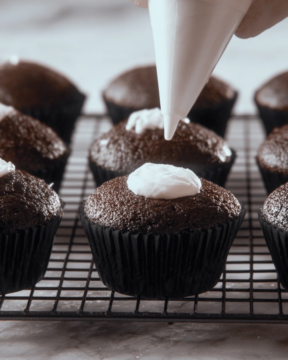 The image shows a close-up of a chocolate cupcake with three layers: the bottom is dark, moist chocolate cake; the middle is a smooth, white creamy filling; and the top is a glossy dark chocolate glaze with a thin white icing swirl decoration. The cupcake has a bite taken out of it, revealing the inside layers. Several similar cupcakes with the same dark chocolate top and white icing swirl are blurred in the background, all sitting on a white marbled surface. photo taken with an iphone --ar 4:5 --v 7