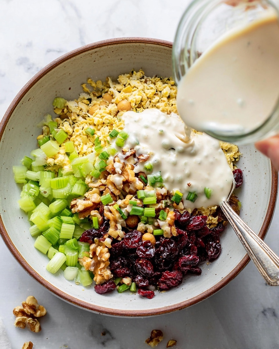 In a white bowl with a brown rim, there is a colorful mix of layers starting with a base of light yellow chickpeas and lentils. On top of this base, there are chopped pale green celery pieces spread around, along with a pile of dark red dried cranberries and light brown chopped walnuts. Green scallions are sprinkled in the middle, adding a fresh bright touch. A creamy white sauce is being poured over the layers from a glass jar by a woman's hand, covering parts of the ingredients. The bowl is on a white marbled surface with some small food crumbs and a silver spoon resting inside the bowl. Photo taken with an iphone --ar 4:5 --v 7