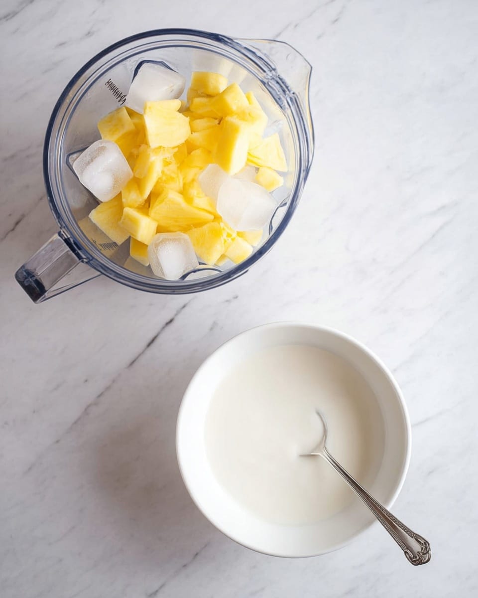 The image shows a clear blender jar filled with large ice cubes and chunks of yellow fruit on top, placed on a white marbled surface. Below the blender jar, there is a white bowl filled with a smooth white liquid, and a silver spoon rests inside the bowl. The scene is bright and clean with a simple arrangement. Photo taken with an iphone --ar 4:5 --v 7