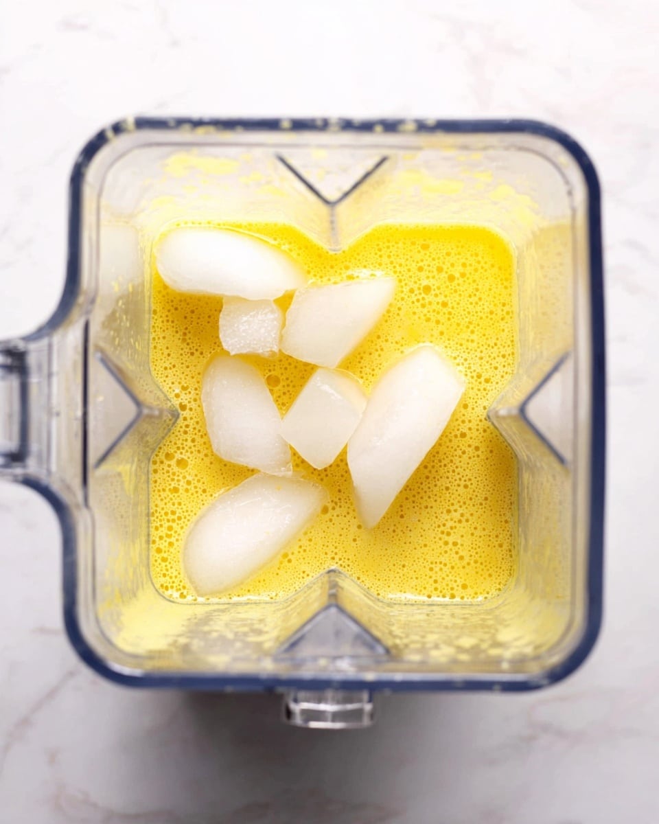 A clear blender container filled with a yellow, slightly frothy liquid at the bottom, topped with several pieces of white ice cubes scattered unevenly over the surface. The blender is placed on a white marbled texture surface, and a metal spoon can be seen on the right side inside the blender. The yellow liquid contrasts against the white ice cubes, showing bubbles and texture within the liquid. photo taken with an iphone --ar 4:5 --v 7