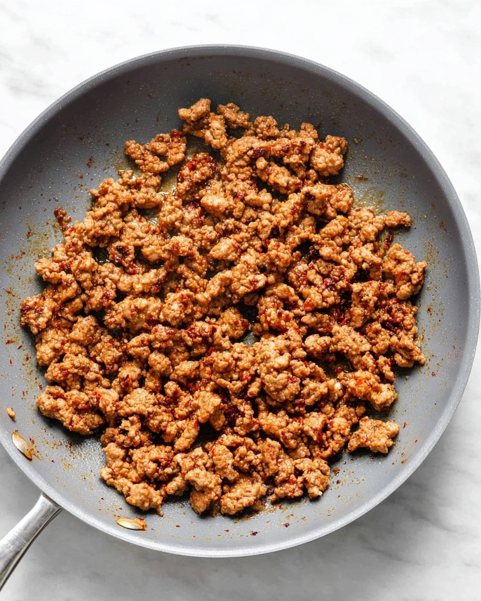 A gray pan filled with cooked ground meat seasoned with spices, showing a mix of small and medium pieces with a browned, slightly crispy texture evenly spread across the pan. The pan rests on a white marbled surface. photo taken with an iphone --ar 4:5 --v 7