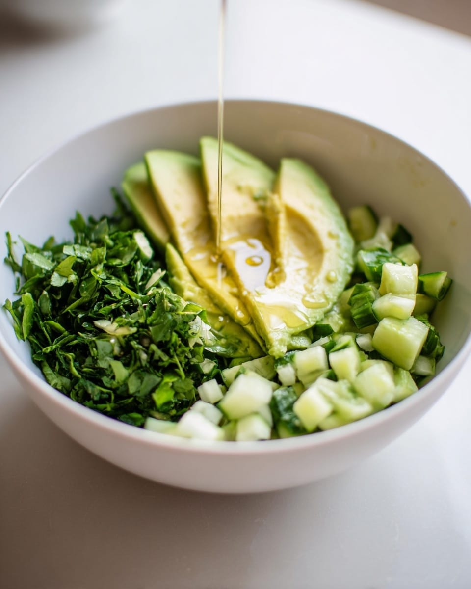 A white bowl sits on a white marbled surface, filled with three main layers: the bottom layer is finely chopped fresh green herbs, the middle layer consists of small diced light green cucumber pieces, and the top layer features thick slices of ripe avocado with light green flesh. A thin stream of golden oil is being poured over the avocado, creating a slight shine and small bubbles on the surface. The background is softly blurred with light tones. photo taken with an iphone --ar 4:5 --v 7