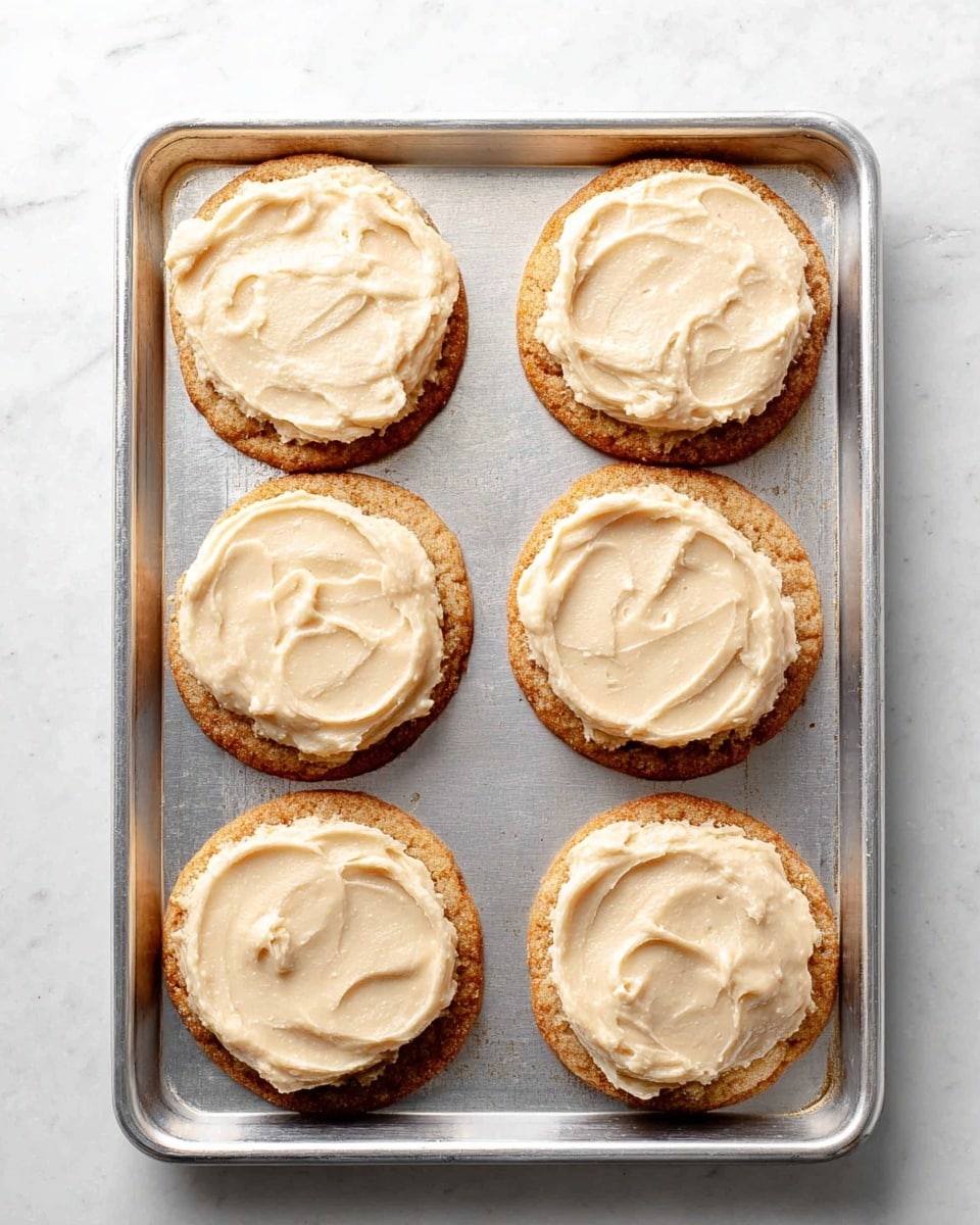 Six round cookies are placed in two rows on a silver baking tray, each cookie topped with a thick, creamy light beige spread evenly covering the surface. The cookies have a golden brown color with a slightly rough texture on the edges. The tray sits on a white marbled surface, creating a clean and bright background. photo taken with an iphone --ar 4:5 --v 7