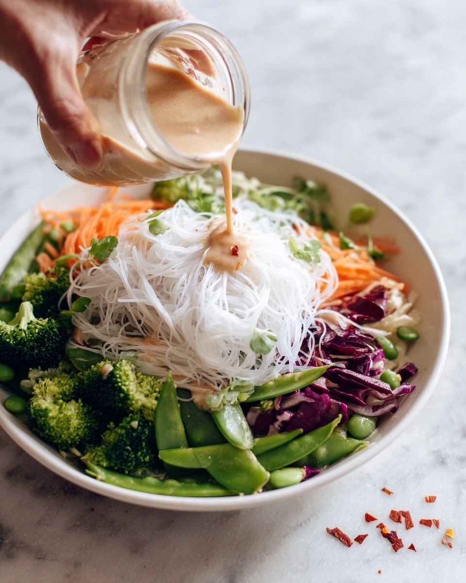 A white bowl holds a colorful salad with three clear layers: the bottom layer is a mix of green broccoli florets, dark green snap peas, purple cabbage, and orange carrot ribbons; the middle layer consists of delicate white rice noodles placed on top of the vegetables; the top layer is a creamy light brown sauce being poured from a small glass jar by a woman's hand above the noodles. The bowl is set on a white marbled surface with a small sprinkle of red flakes nearby. photo taken with an iphone --ar 4:5 --v 7