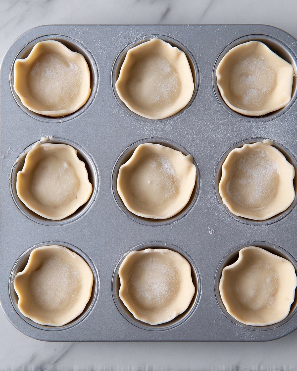 The image shows a gray muffin tray with twelve cups, each lined with a thin layer of pale, uncooked dough pressed into the shape of the cups. The dough edges are crimped gently, creating soft folds along the top rim of each cup. The surface of the dough looks smooth and slightly dusted with flour, and the tray sits on a white marbled surface. photo taken with an iphone --ar 4:5 --v 7