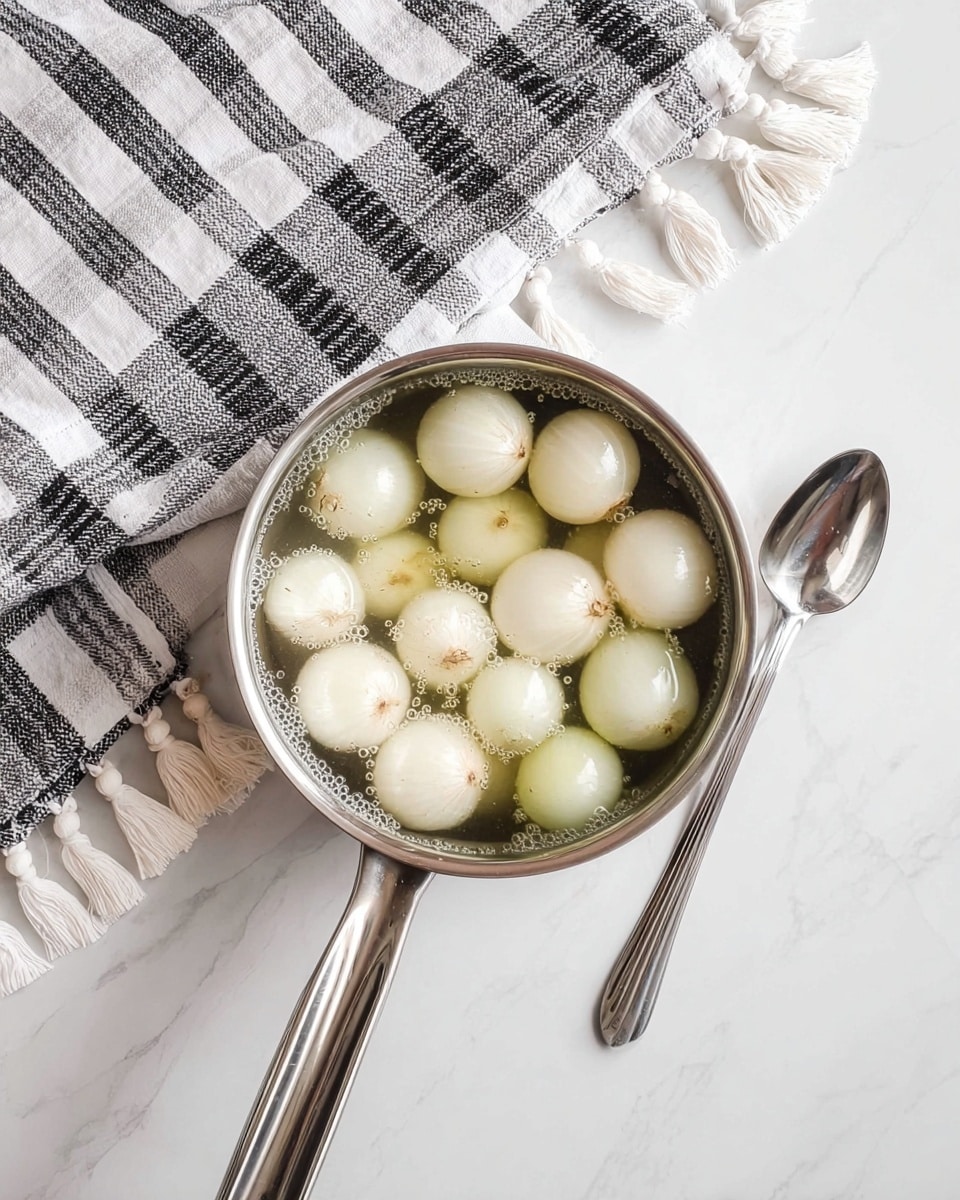 A small silver pot filled with about twenty small white onions submerged in clear water, showing slight bubbles on the onion surfaces, resting on a white marbled surface with a black and white checkered cloth with white tassels gathered to the left side, and a shiny silver spoon placed on the right side, the pot handle extending towards the bottom center of the image. photo taken with an iphone --ar 4:5 --v 7