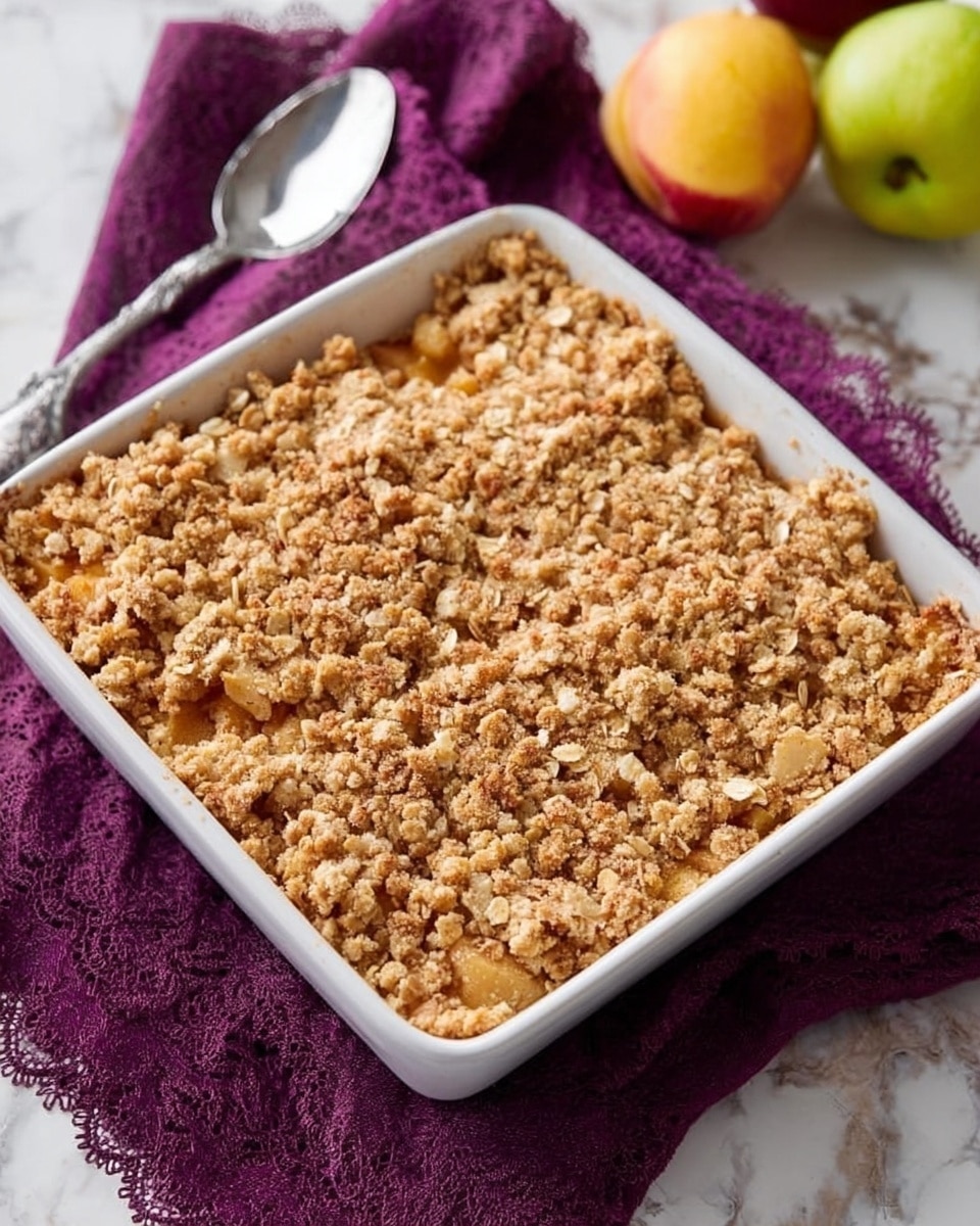 A square white baking dish filled with a golden-brown crumble topping that has a rough, crumbly texture, with visible oats and small clusters scattered evenly across the surface. The dish sits on a deep purple cloth with lace edges, placed on a white marbled surface, with a silver spoon resting nearby. Two fruits, one yellow-green and one reddish yellow, partially visible near the top edge, add a touch of color to the scene. Photo taken with an iphone --ar 4:5 --v 7