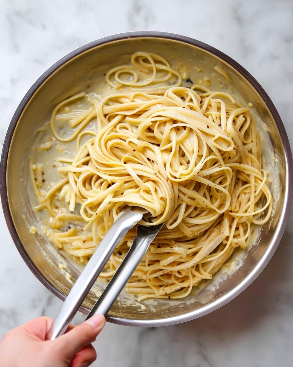 A close-up top view of a silver pan filled with creamy pale yellow pasta noodles coated in a light sauce with small bits of seasoning mixed throughout. The noodles are long and flat, gathered in the center with silver tongs lifting a portion. A woman's hand holds the tongs, gripping them from the bottom left corner. The sauce clings lightly to the noodles and pan, showing a smooth and slightly thick texture. The background is a white marbled surface. photo taken with an iphone --ar 4:5 --v 7