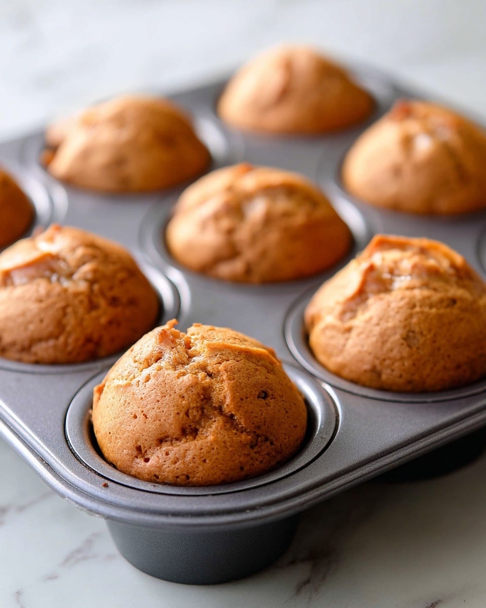 The image shows a close-up of six light brown muffins inside a dark gray muffin tin, each muffin rising slightly above the edge of each round mold. The muffins have a smooth but slightly cracked texture on the top, showing a soft baked surface. The muffin tin sits on a white marbled surface with soft lighting highlighting the golden brown color of the muffins and the metallic sheen of the tin. photo taken with an iphone --ar 4:5 --v 7