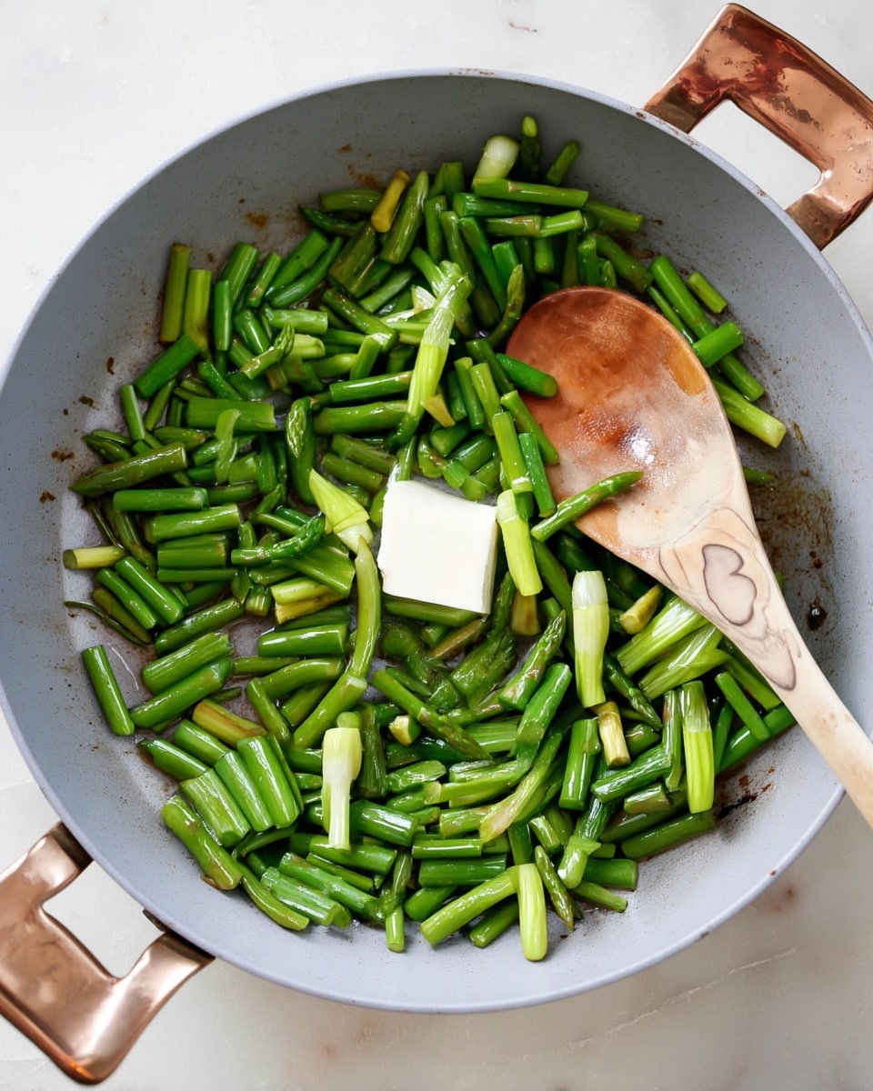 A light gray pan filled with many small, bright green chopped asparagus pieces evenly spread out. In the center of the pan, there is a small square of white butter starting to melt. A wooden spoon with a smooth texture and brown patterns rests on the right side of the pan, mixing the asparagus. The pan has two copper-colored handles visible on the left side. The background is a white marbled surface. photo taken with an iphone --ar 4:5 --v 7