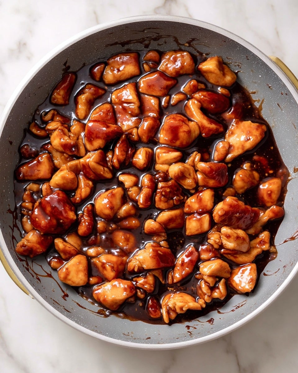 A large grey pan filled with many small pieces of chicken coated in a thick, shiny dark brown sauce, evenly spread across the pan's center. The chicken pieces have a rich, glossy texture, showing a smooth surface with some darker spots from the sauce. The pan rests on a white marbled surface with the edges splattered lightly by the sauce, giving the dish a freshly cooked, saucy look. photo taken with an iphone --ar 4:5 --v 7