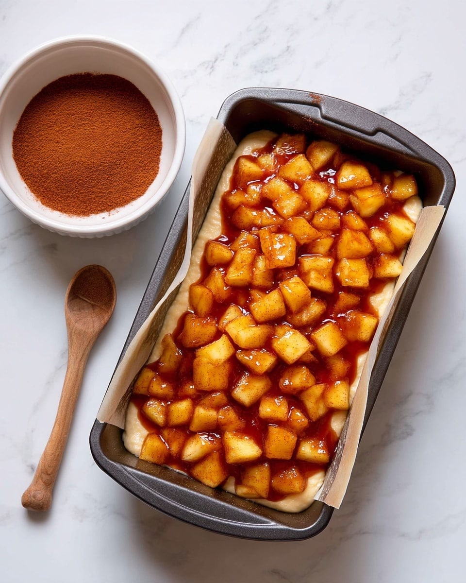 A rectangular dark metal baking pan lined with parchment paper holds a single layer of thick, creamy beige batter on the bottom, topped with an even layer of small, diced yellow apple pieces coated lightly in a reddish-brown cinnamon sauce. Next to the pan is a white bowl filled with a reddish-brown powder and a small wooden spoon resting inside. The scene is set on a white marbled surface. photo taken with an iphone --ar 4:5 --v 7
