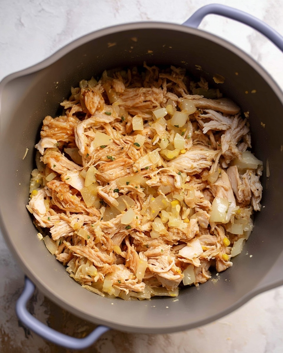 Inside a grey cooking pot, there is a mixture of shredded light brown cooked meat layered with small, soft, translucent chopped onions spread evenly throughout. The meat fibers are visible, and the onions add a slight shine and pale yellow tint to the dish. The pot is placed on a white marbled surface. Photo taken with an iphone --ar 4:5 --v 7