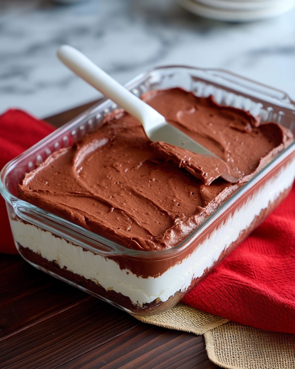 The image shows a clear glass rectangular dish with two thick layers of dessert. The bottom layer is white, smooth, and creamy, while the top layer is a dark brown, rich chocolate spread in a thick, textured way. A woman's hand is using a white spatula to spread the chocolate layer evenly across the dish. The dish sits on a dark wooden surface with a burlap piece and a red cloth nearby, but the background should be imagined as white marbled texture. Photo taken with an iphone --ar 4:5 --v 7