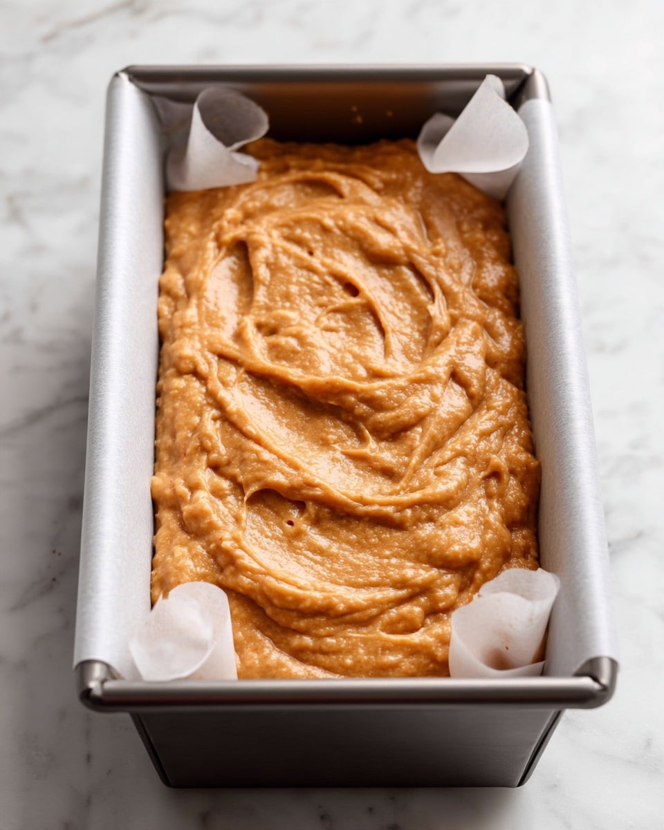 A close-up view of a rectangular metal baking pan lined with white parchment paper, filled with smooth, thick, light brown batter evenly spread inside it. The batter has a slightly textured surface with visible swirls and soft peaks, giving it a moist and rich look. The pan is placed on a white marbled surface that adds a clean and bright touch to the image. photo taken with an iphone --ar 4:5 --v 7